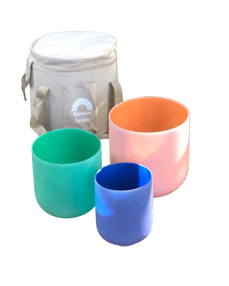 Set of colorful tibetan singing bowls on a woven mat with a cooler labeled 'Rainbow Sounds' in the background.