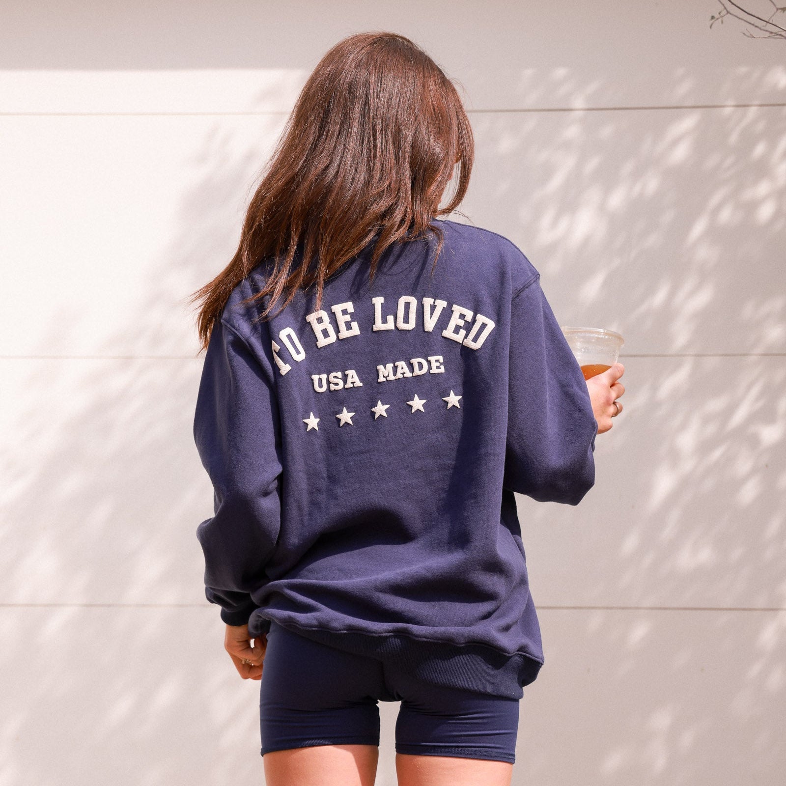 Person wearing a navy blue sweatshirt with text, holding a drink against a light background