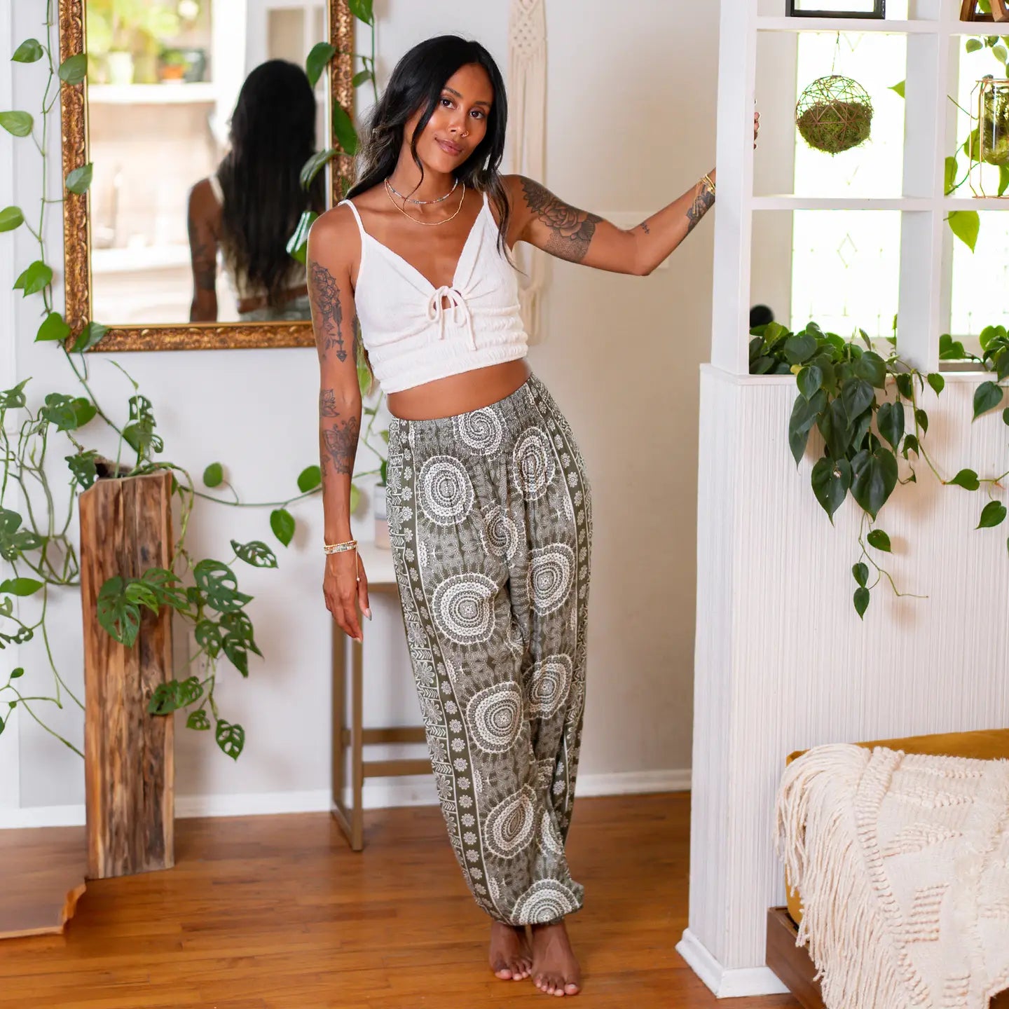 Woman in a white top and patterned pants standing in a room with plants and a mirror.