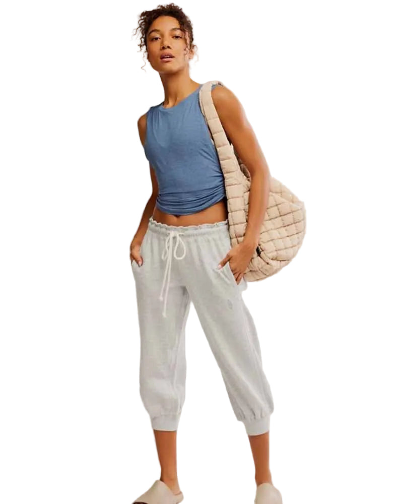 Woman wearing a blue sleeveless top and light gray sweatpants, holding a beige bag on a white background