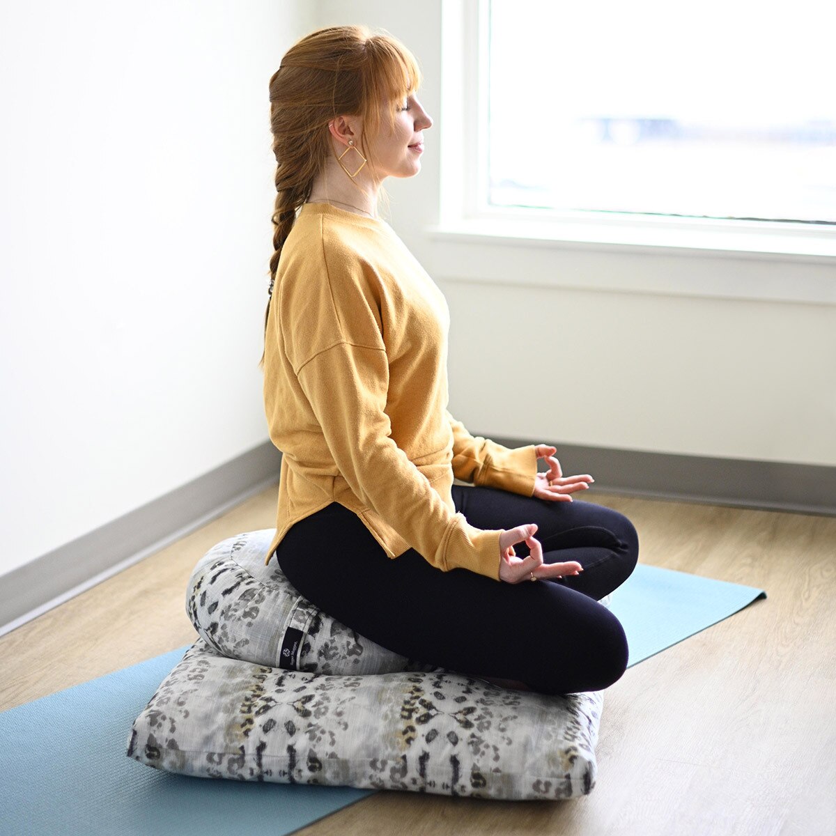 Woman meditating on a cushion in a bright room with large windows.