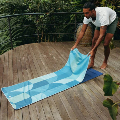 Man unfolding a blue patterned towel on a wooden deck with greenery in the background