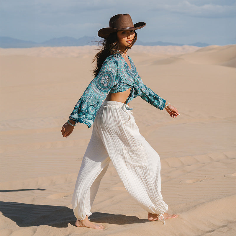 Woman in a patterned top and white pants walking on sand dunes with mountains in the background