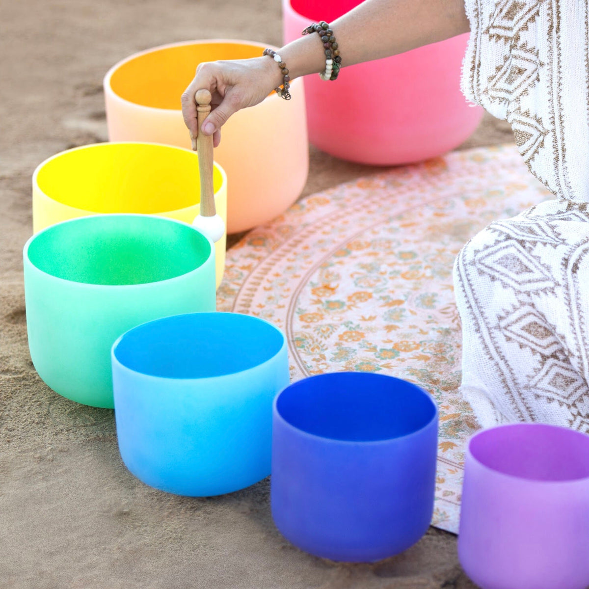 Colorful singing bowls on a sandy surface with a person holding a mallet.