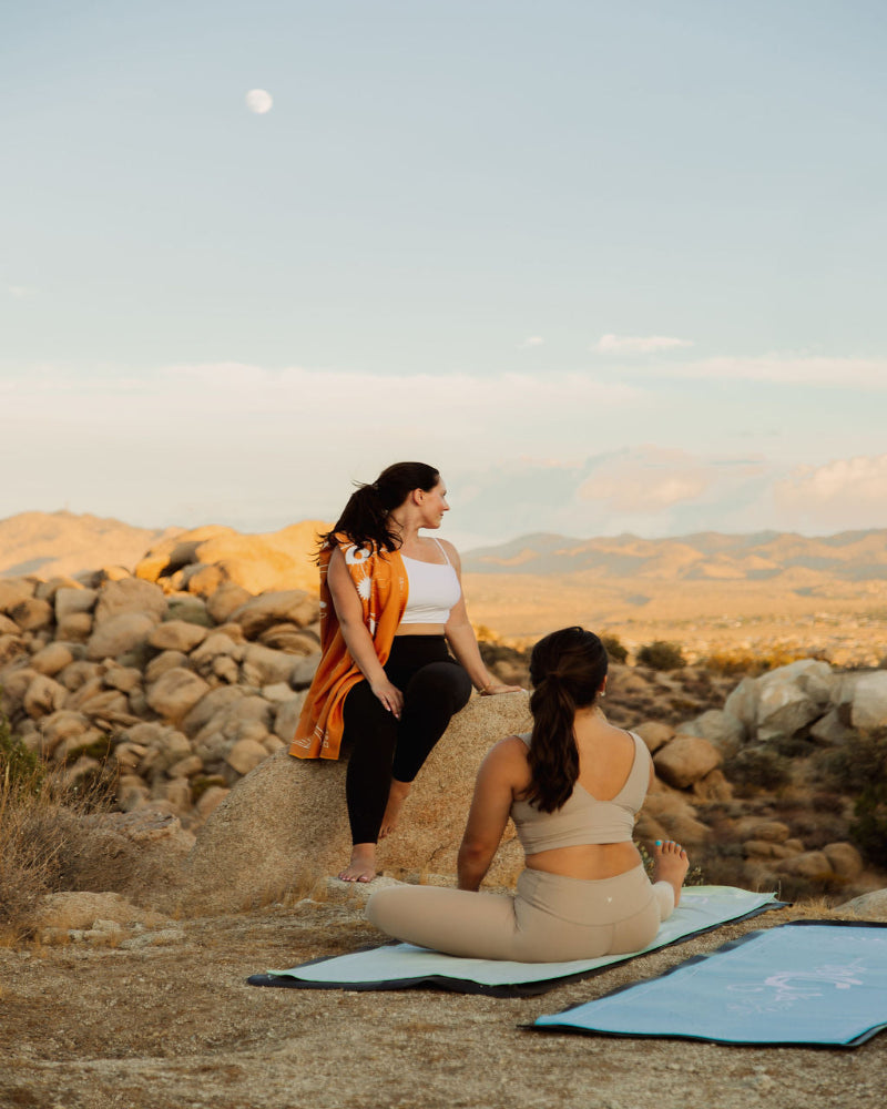 Two women practicing yoga in a desert landscape with mountains and a clear sky.