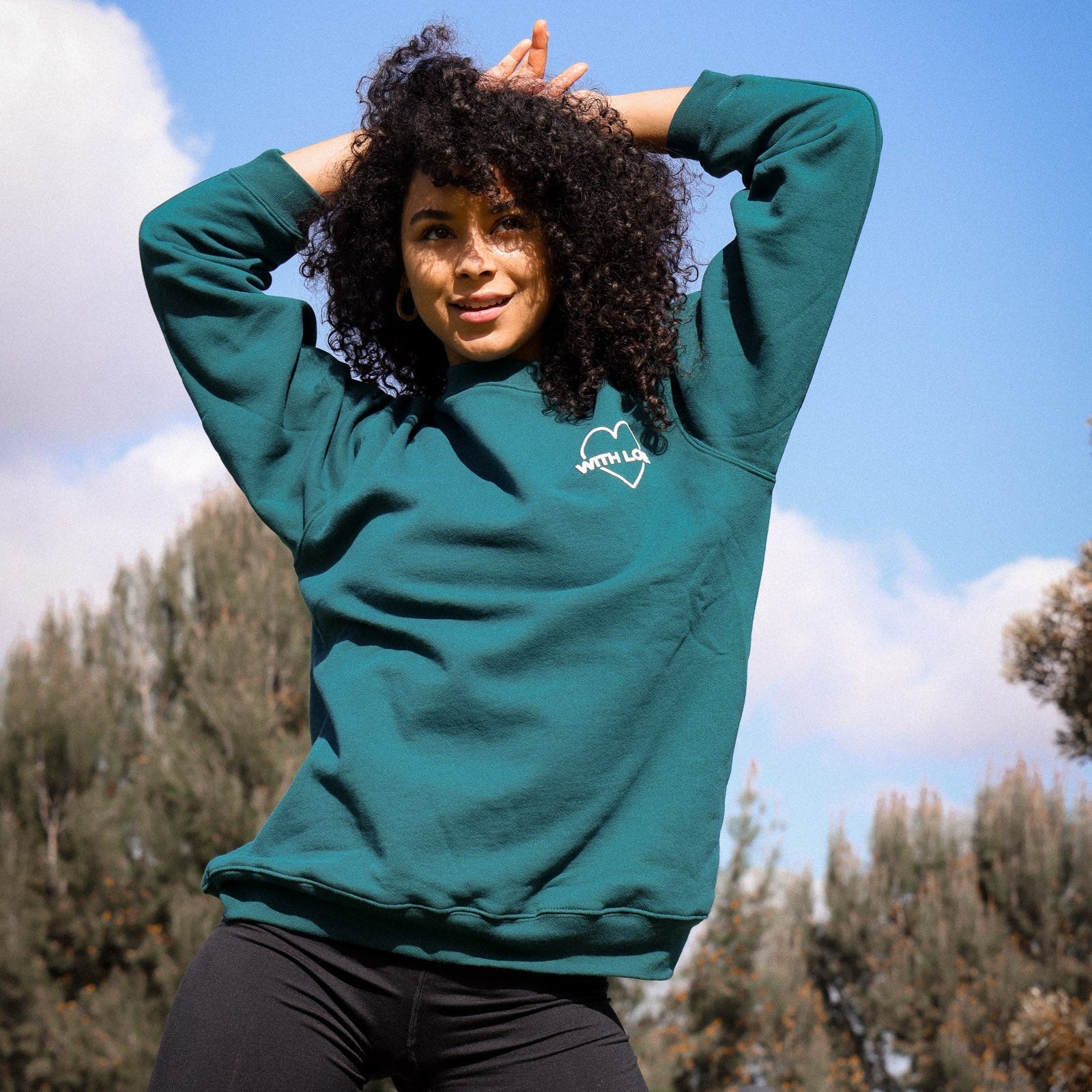 Person wearing a green sweatshirt with a logo outdoors against a blue sky.