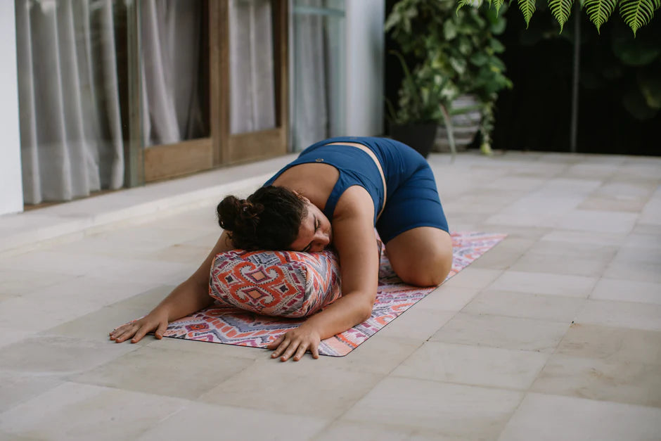 Woman practicing yoga on a mat with a pillow under her head in an outdoor setting.