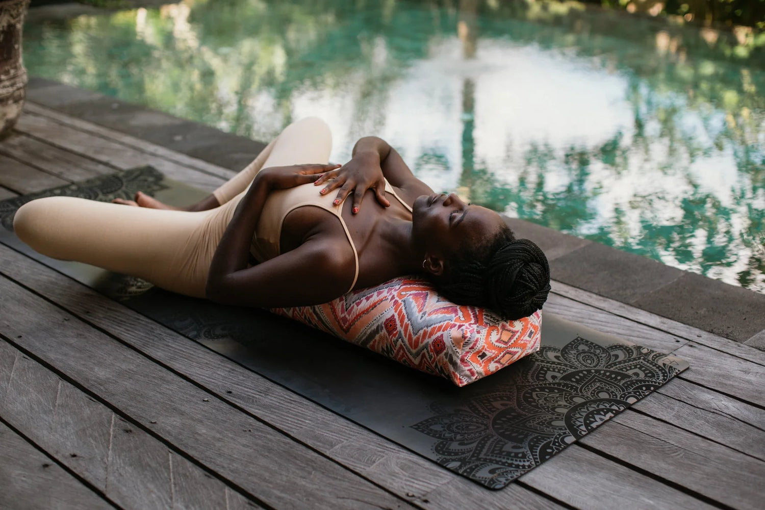 A woman in beige activewear lies on a patterned yoga pillow on a black yoga mat, positioned on a wooden deck next to a swimming pool.