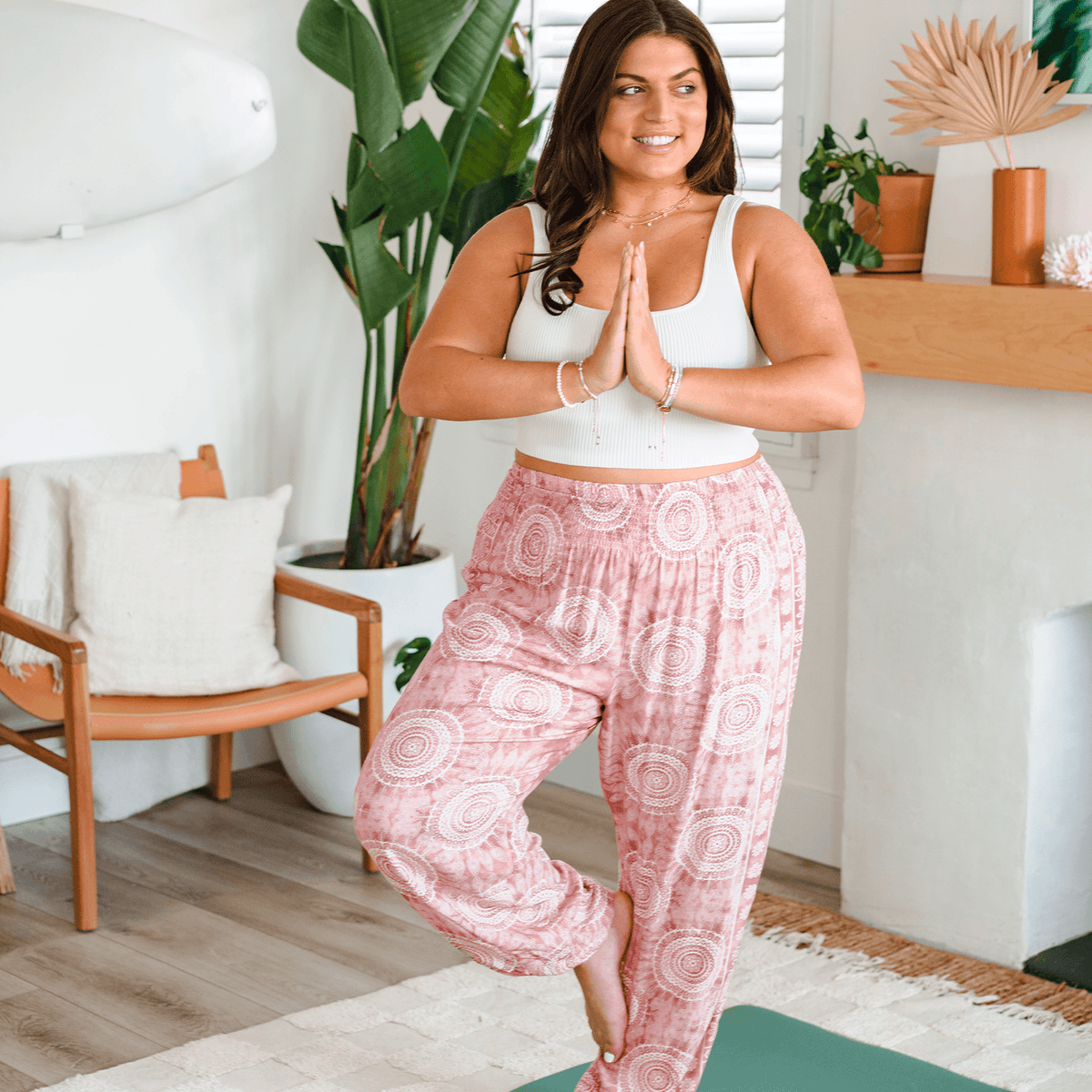 Woman in pink patterned pants and white top standing on a yoga mat in a home setting.