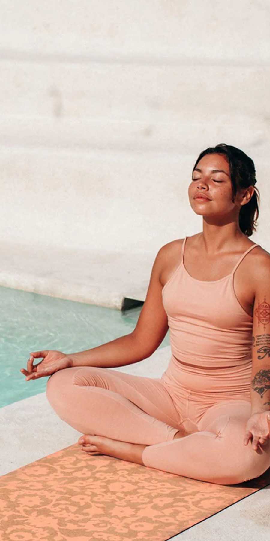 Woman in peach swimsuit meditating on a mat by a pool.