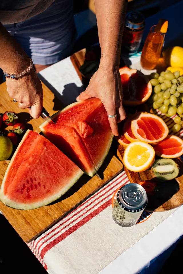 Person cutting watermelon on a wooden cutting board with fruits and drinks in the background.
