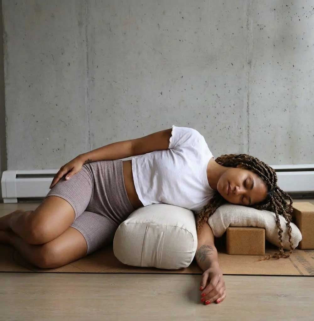 Person lying on a yoga mat with pillows and blocks in a minimalistic room.