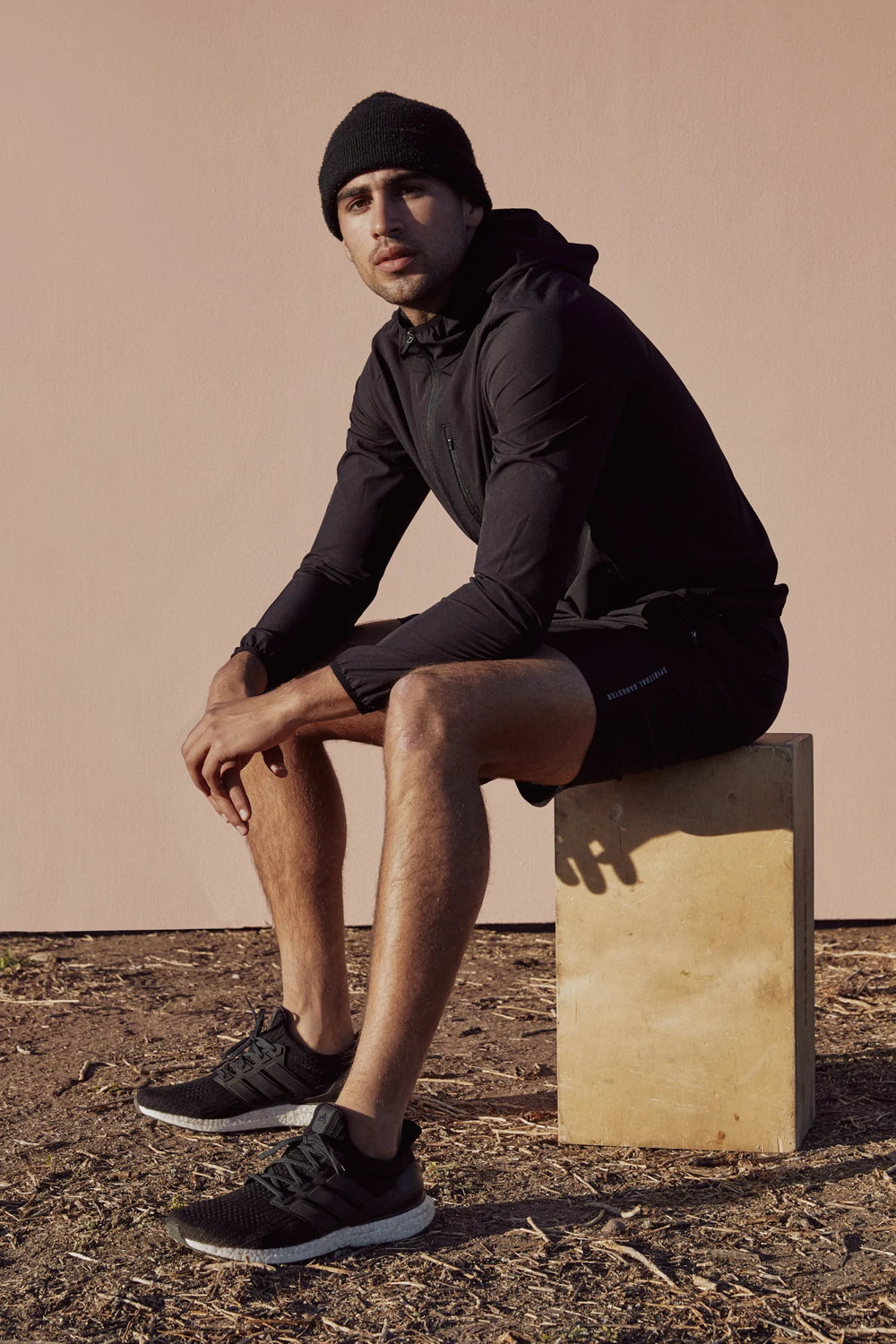 Man in black athletic wear sitting on a block against a beige wall.