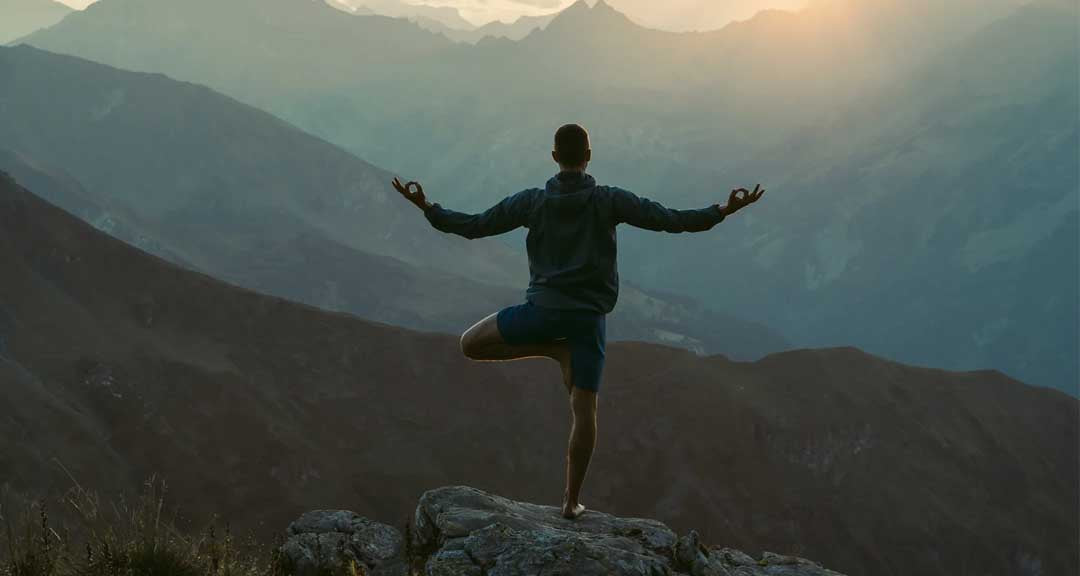Person practicing yoga on a mountain top with a scenic view of mountains and sunset.