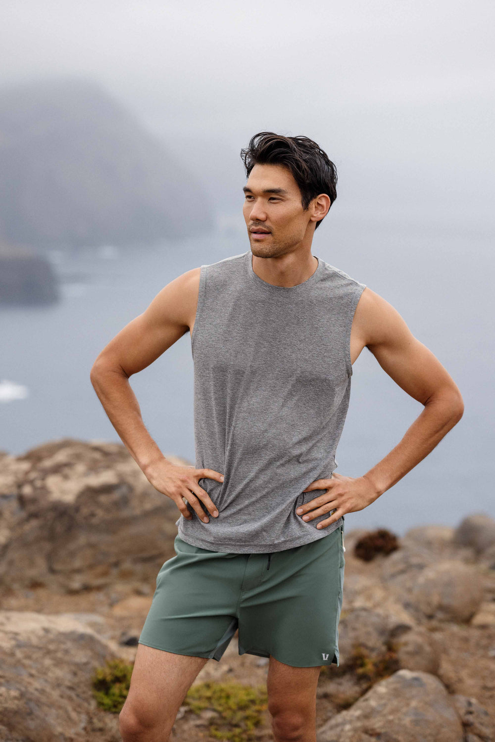 Man wearing a gray sleeveless shirt and green shorts standing on rocky terrain with a misty background.