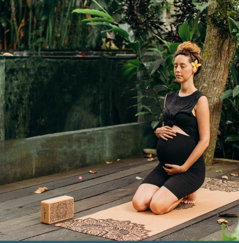 Pregnant woman practicing yoga outdoors with a yoga block on a mat.