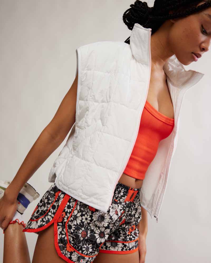 Woman wearing a white puffer vest, orange top, and patterned shorts on a plain background